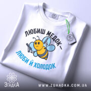 Футболка медок холодок, біла, складена на поверхні, фон світло-сірий, принт з бджолою, яка тримає пістолети, текст чорного та блакитного кольору, з етикеткою на рукаві.