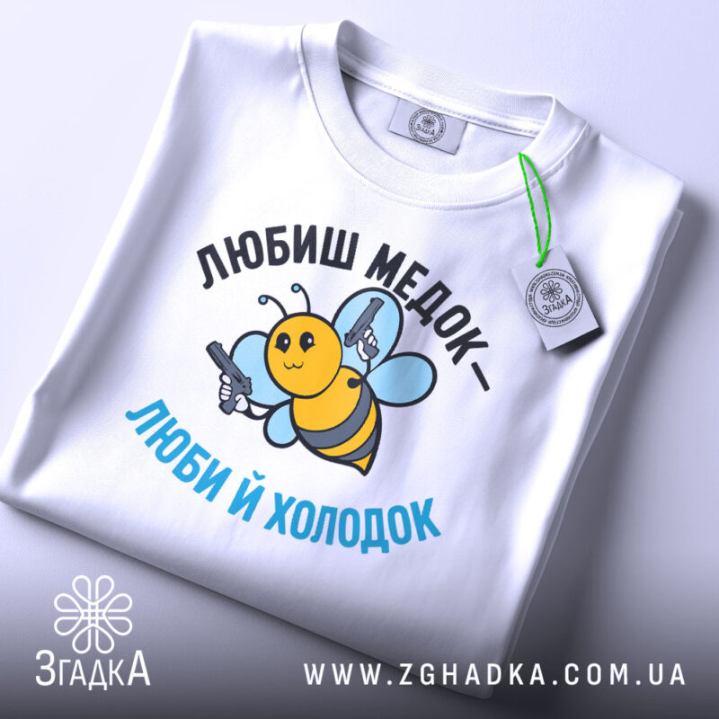 Футболка медок холодок, біла, складена на поверхні, фон світло-сірий, принт з бджолою, яка тримає пістолети, текст чорного та блакитного кольору, з етикеткою на рукаві.