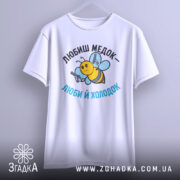 Футболка медок холодок, біла, підвішена на вішалці, фон світло-сірий, принт з бджолою, яка тримає пістолети, текст чорного та блакитного кольору.