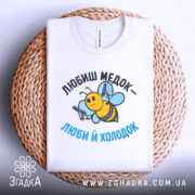 Футболка медок холодок, біла, складена на плетеній основі, фон світло-сірий, принт з бджолою, яка тримає пістолети, текст чорного та блакитного кольору.