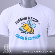 Футболка медок холодок, біла, складена на поверхні, фон світло-сірий, принт з бджолою, яка тримає пістолети, текст чорного та блакитного кольору, з етикеткою на рукаві.