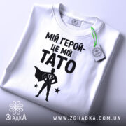 Футболка мій герой тато, біла, складена на столі, фон світло-сірий, принт чорний з написом 'МІЙ ГЕРОЙ-ЦЕ МІЙ TATO' та силуетом супергероя, з зеленим ярликом.