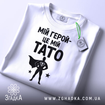 Футболка мій герой тато, біла, складена на столі, фон світло-сірий, принт чорний з написом 'МІЙ ГЕРОЙ-ЦЕ МІЙ TATO' та силуетом супергероя, з зеленим ярликом.
