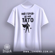 Футболка мій герой тато, біла, підвішена на вішалці, фон світло-сірий, принт чорний з написом 'МІЙ ГЕРОЙ-ЦЕ МІЙ TATO' та силуетом супергероя.