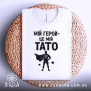 Футболка мій герой тато, біла, складена на плетеній кошику, фон світло-сірий, принт чорний з написом 'МІЙ ГЕРОЙ-ЦЕ МІЙ TATO' та силуетом супергероя.