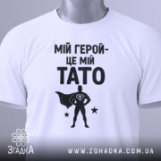 Футболка мій герой тато, біла, складена на столі, фон світло-сірий, принт чорний з написом 'МІЙ ГЕРОЙ-ЦЕ МІЙ TATO' та силуетом супергероя, з зеленим ярликом.