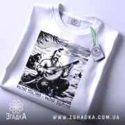 Футболка моя пісня зброя, складена біла футболка, ракурс зверху, фон світло-сірий, на футболці принт з кобзарем, який грає на бандурі, з зеленим ярликом.