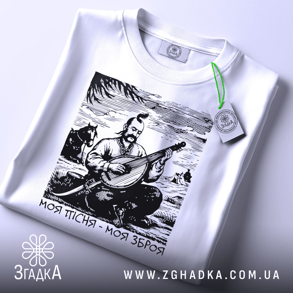 Футболка моя пісня зброя, складена біла футболка, ракурс зверху, фон світло-сірий, на футболці принт з кобзарем, який грає на бандурі, з зеленим ярликом.