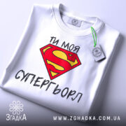 Футболка моя супергьорл, біла, складена на поверхні, фон світло-сірий, принт з червоним логотипом Супермена та текстом "ТИ МОЯ СУПЕРГЬОРЛ" чорного кольору, з етикеткою на зеленому шнурку.