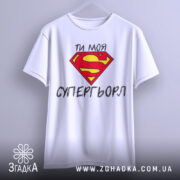 Футболка моя супергьорл, біла, підвішена на вішалці, фон світло-сірий, принт з червоним логотипом Супермена та текстом "ТИ МОЯ СУПЕРГЬОРЛ" чорного кольору.