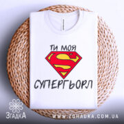 Футболка моя супергьорл, біла, складена на круглому плетеному кошику, фон світло-сірий, принт з червоним логотипом Супермена та текстом "ТИ МОЯ СУПЕРГЬОРЛ" чорного кольору.