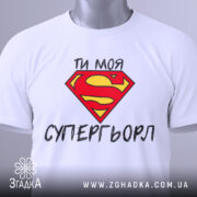 Футболка моя супергьорл, біла, складена на поверхні, фон світло-сірий, принт з червоним логотипом Супермена та текстом "ТИ МОЯ СУПЕРГЬОРЛ" чорного кольору, з етикеткою на зеленому шнурку.