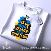 Футболка моя земля фортеця, біла, складена на столі, фон світло-сірий, принт з написом 'Моя земля - моя фортеця' у жовто-синіх кольорах, з зеленим ярликом.