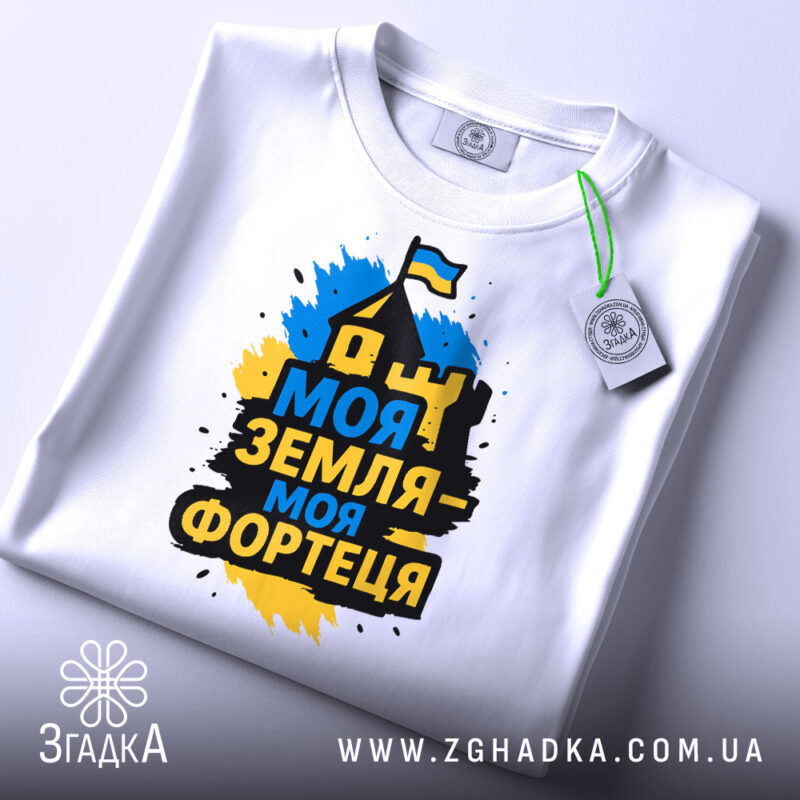 Футболка моя земля фортеця, біла, складена на столі, фон світло-сірий, принт з написом 'Моя земля - моя фортеця' у жовто-синіх кольорах, з зеленим ярликом.