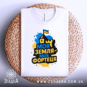 Футболка моя земля фортеця, біла, складена на плетеному кошику, фон світло-сірий, принт з написом 'Моя земля - моя фортеця' у жовто-синіх кольорах, видима частина горловини.