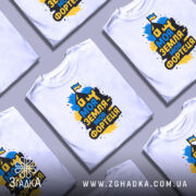 Футболка моя земля фортеця, біла, зображена на манекені з правого боку, фон світло-сірий, принт з написом 'Моя земля - моя фортеця' у жовто-синіх кольорах з зображенням будинку та прапора.