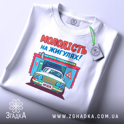 Футболка молодість на жигулях, біла, складена на столі, фон світло-сірий, принт з автомобілем та написом 'МОЛОДІСТЬ НА ЖИГУЛЯХ!' яскравий, з елементами дизайну.