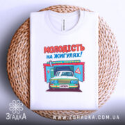 Футболка молодість на жигулях, біла, складена на плетеному кошику, фон світлий, принт з автомобілем та написом 'МОЛОДІСТЬ НА ЖИГУЛЯХ!' яскравий, з чіткими кольоровими елементами.