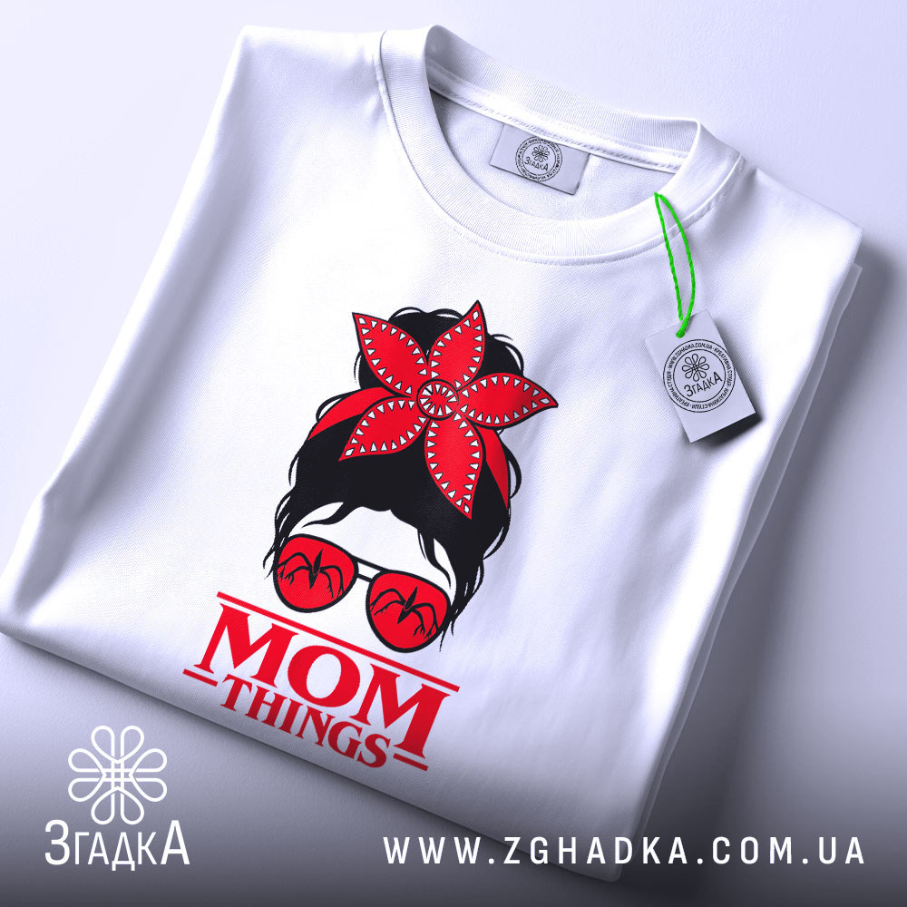 Футболка Mom Thing, білого кольору, зображена зверху, на фоні світлого кольору, принт з червоною квіткою на голові жінки в окулярах та написом 'MOM THINGS' під малюнком, з етикеткою на комірці.