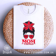 Футболка Mom Thing, білого кольору, зображена зверху на плетеному кошику, фон світлий, принт з червоною квіткою на голові жінки в окулярах та написом 'MOM THINGS' під малюнком.
