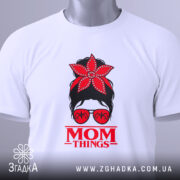 Футболка Mom Thing, білого кольору, зображена зверху, на фоні світлого кольору, принт з червоною квіткою на голові жінки в окулярах та написом 'MOM THINGS' під малюнком, з етикеткою на комірці.