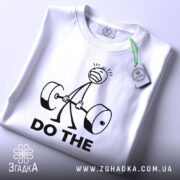 Футболка мотивація для залу, біла, зображена зверху, на ній видно етикетку з зеленим шнурком, фон світло-сірий, принт з малюнком людини, що піднімає штангу, та текстом 'DO THE' чорного кольору.
