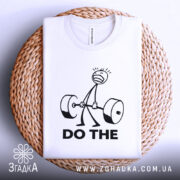 Футболка мотивація для залу, біла, зображена зверху на плетеній кошику, фон світло-сірий, принт з малюнком людини, що піднімає штангу, та текстом 'DO THE' чорного кольору.