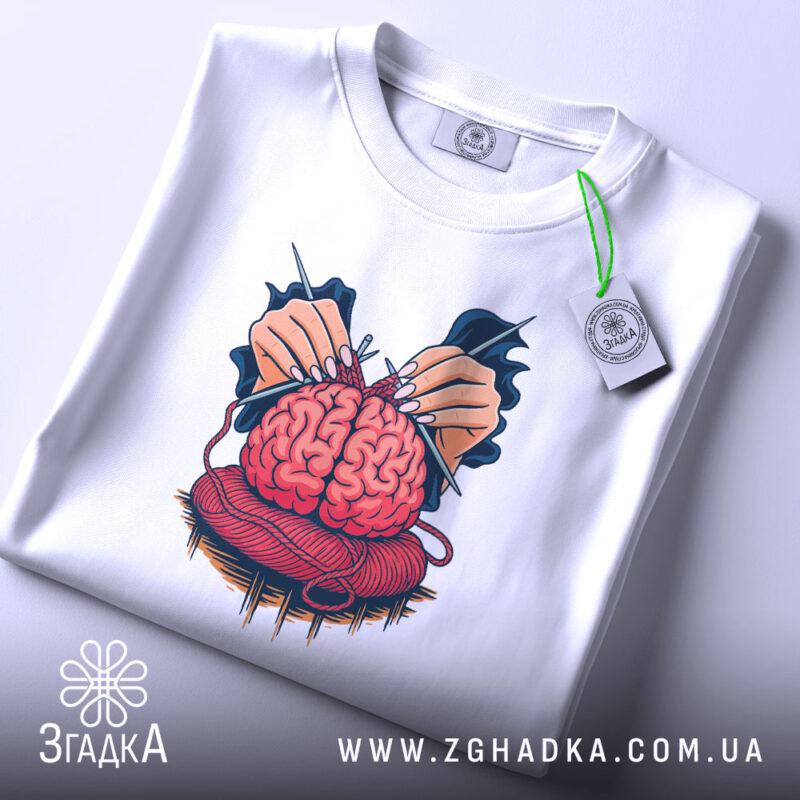 Футболка мозок в клубку, біла, зображена з верхнього ракурсу, на світлому фоні, принт з червоним мозком, що оточений руками з голками, з етикеткою на шнурку.