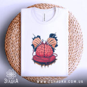 Футболка мозок в клубку, біла, зображена зверху на плетеній основі, на світлому фоні, принт з червоним мозком, що оточений руками з голками та клубком червоної пряжі.