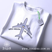 Футболка мрія не вмирає, біла, складена на столі, фон світло-сірий, принт з літаком та написом 'МРІЯ НІКОЛИ НЕ ВМИРАЄ' у чорному кольорі з жовтим сердечком, на комірці видна етикетка.