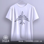 Футболка мрія не вмирає, біла, підвішена на вішалці, фон світло-сірий, принт з літаком та написом 'МРІЯ НІКОЛИ НЕ ВМИРАЄ' у чорному кольорі з жовтим сердечком.