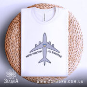 Футболка мрія не вмирає, біла, складена на кошику, фон світло-сірий, принт з літаком та написом 'МРІЯ НІКОЛИ НЕ ВМИРАЄ' у чорному кольорі з жовтим сердечком, видна етикетка на комірці.