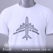 Футболка мрія не вмирає, біла, складена на столі, фон світло-сірий, принт з літаком та написом 'МРІЯ НІКОЛИ НЕ ВМИРАЄ' у чорному кольорі з жовтим сердечком, на комірці видна етикетка.