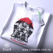 Футболка музика і залізо, біла, складена на столі, фон світлий, принт зображає м'язистого чоловіка на тренажері, червоний фон з написом 'ДВІ РАДОСТІ – МУЗИКА І ЗАЛІЗО', на комірці є етикетка.