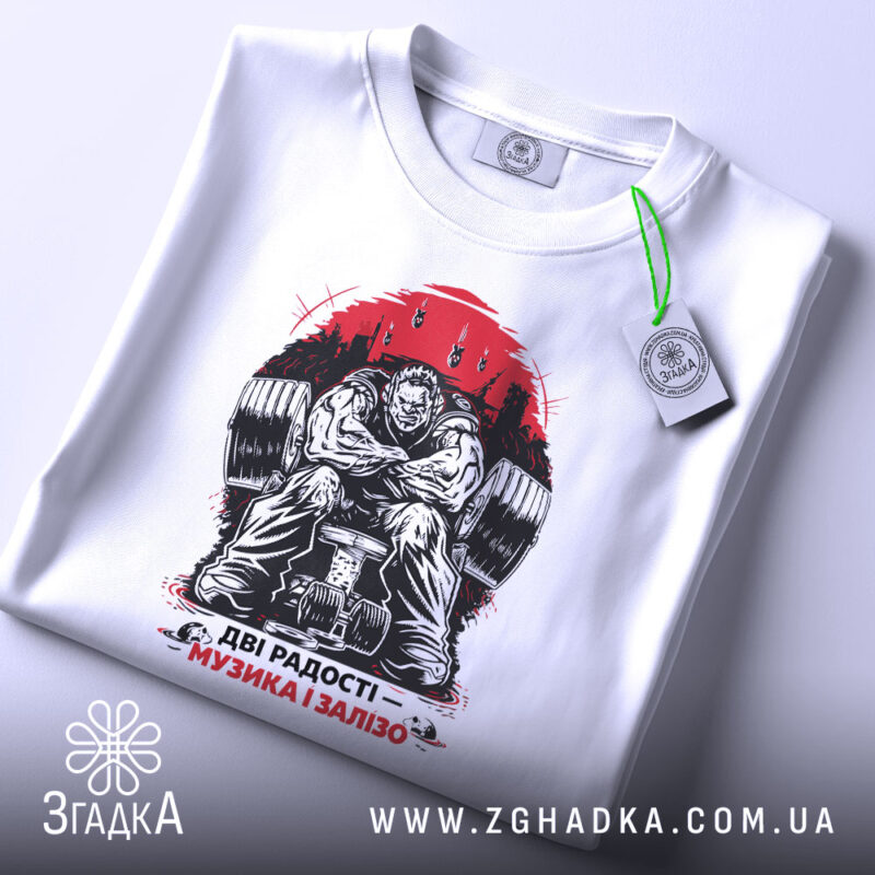 Футболка музика і залізо, біла, складена на столі, фон світлий, принт зображає м'язистого чоловіка на тренажері, червоний фон з написом 'ДВІ РАДОСТІ – МУЗИКА І ЗАЛІЗО', на комірці є етикетка.