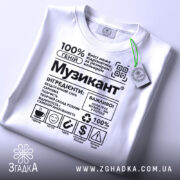 Футболка музикант білого кольору, складена на поверхні, з принтом у чорному кольорі, на фоні світло-сірого кольору, з зеленим ярликом, що звисає з коміра.