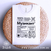 Футболка музикант білого кольору, складена на плетеній кошику, з принтом у чорному кольорі, на фоні світло-сірого кольору, деталі принта добре видно.
