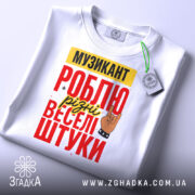 Футболка музикант білого кольору, складена на поверхні, фон світлий, принт з текстом "МУЗИКАНТ РОБЛЮ різні веселі штуки" в червоному, жовтому та чорному кольорах, з малюнком руки в жесті рок-знака, на футболці є етикетка.