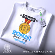 Футболка музикант, біла, зображена зверху, на ній підвішена етикетка, фон світло-сірий, принт з медаллю та текстом "ГАРНИЙ МУЗИКАНТ знає все І навіть більше..." у яскравих кольорах.