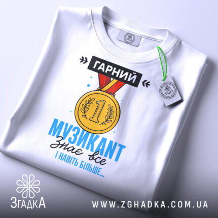 Футболка музикант, біла, зображена зверху, на ній підвішена етикетка, фон світло-сірий, принт з медаллю та текстом "ГАРНИЙ МУЗИКАНТ знає все І навіть більше..." у яскравих кольорах.