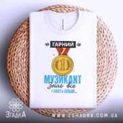 Футболка музикант, біла, зображена зверху на плетеній кошику, фон світло-сірий, принт з медаллю та текстом "ГАРНИЙ МУЗИКАНТ знає все І навіть більше..." у яскравих кольорах.