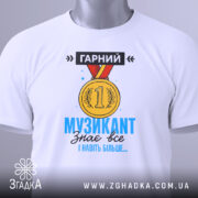 Футболка музикант, біла, зображена зверху, на ній підвішена етикетка, фон світло-сірий, принт з медаллю та текстом "ГАРНИЙ МУЗИКАНТ знає все І навіть більше..." у яскравих кольорах.