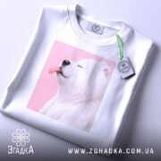 Футболка милий білий ведмедик, біла, зображена зверху на столі, фон світло-сірий, принт ведмедика з висунутим язиком на рожевому фоні, з етикеткою на комірці.