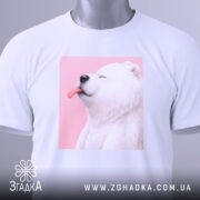 Футболка милий білий ведмедик, біла, зображена зверху на столі, фон світло-сірий, принт ведмедика з висунутим язиком на рожевому фоні, з етикеткою на комірці.