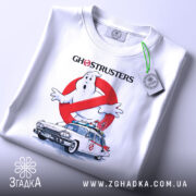 Футболка мисливці за привидами, біла, складена на поверхні, показана з верхнього ракурсу, фон світло-сірий, принт з зображенням привида та автомобіля з фільму, з написом 'GHOSTRUSTERS', на футболці є етикетка з зеленим шнурком.