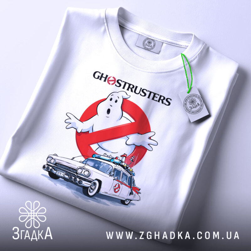 Футболка мисливці за привидами, біла, складена на поверхні, показана з верхнього ракурсу, фон світло-сірий, принт з зображенням привида та автомобіля з фільму, з написом 'GHOSTRUSTERS', на футболці є етикетка з зеленим шнурком.