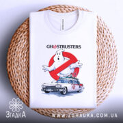 Футболка мисливці за привидами, біла, складена на плетеній кошику, показана з верхнього ракурсу, фон світло-сірий, принт з зображенням привида та автомобіля з фільму, з написом 'GHOSTRUSTERS', видима етикетка на горловині.