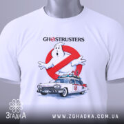 Футболка мисливці за привидами, біла, складена на поверхні, показана з верхнього ракурсу, фон світло-сірий, принт з зображенням привида та автомобіля з фільму, з написом 'GHOSTRUSTERS', на футболці є етикетка з зеленим шнурком.