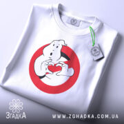 Футболка мисливці за привидами, біла, складена на поверхні, фон світло-сірий, принт з привидом, що тримає червоне сердечко в руках, з етикеткою на зеленій стрічці.