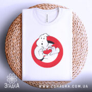 Футболка мисливці за привидами, біла, складена на плетеній кошику, фон світло-сірий, принт з привидом, що тримає червоне сердечко в руках, з видимим ярликом.